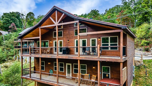 A two-story wooden cabin with multiple balconies, outdoor seating, and a scenic hillside view. - New ModernRustic House in Smokys-4BR/Pools/Hot Tub/Billards/Games (Gatlinburg)
