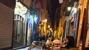 Une personne tenant un verre de vin dans une rue étroite, bordée de bâtiments des deux côtés.