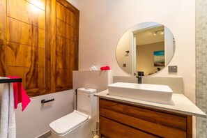 A bathroom with a wooden panel wall, a white toilet, a white sink with a mirror above it, and a tiled floor. - Brand New two Bedrooms Apartment at One Bay Residence (Grand-Baie)