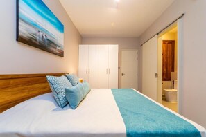 A bedroom with a bed, a painting, a wardrobe, and a bathroom visible through an open door. - Brand New two Bedrooms Apartment at One Bay Residence (Grand-Baie)
