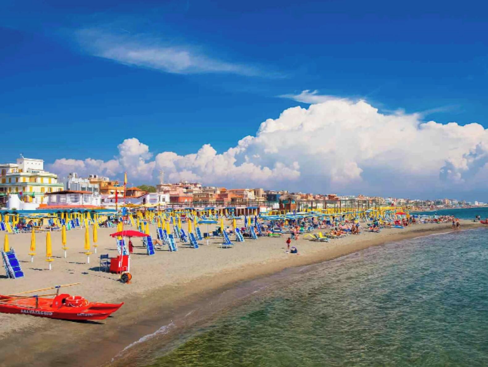 Una playa con hileras de sombrillas y tumbonas, una torre de salvavidas y un bote rojo.