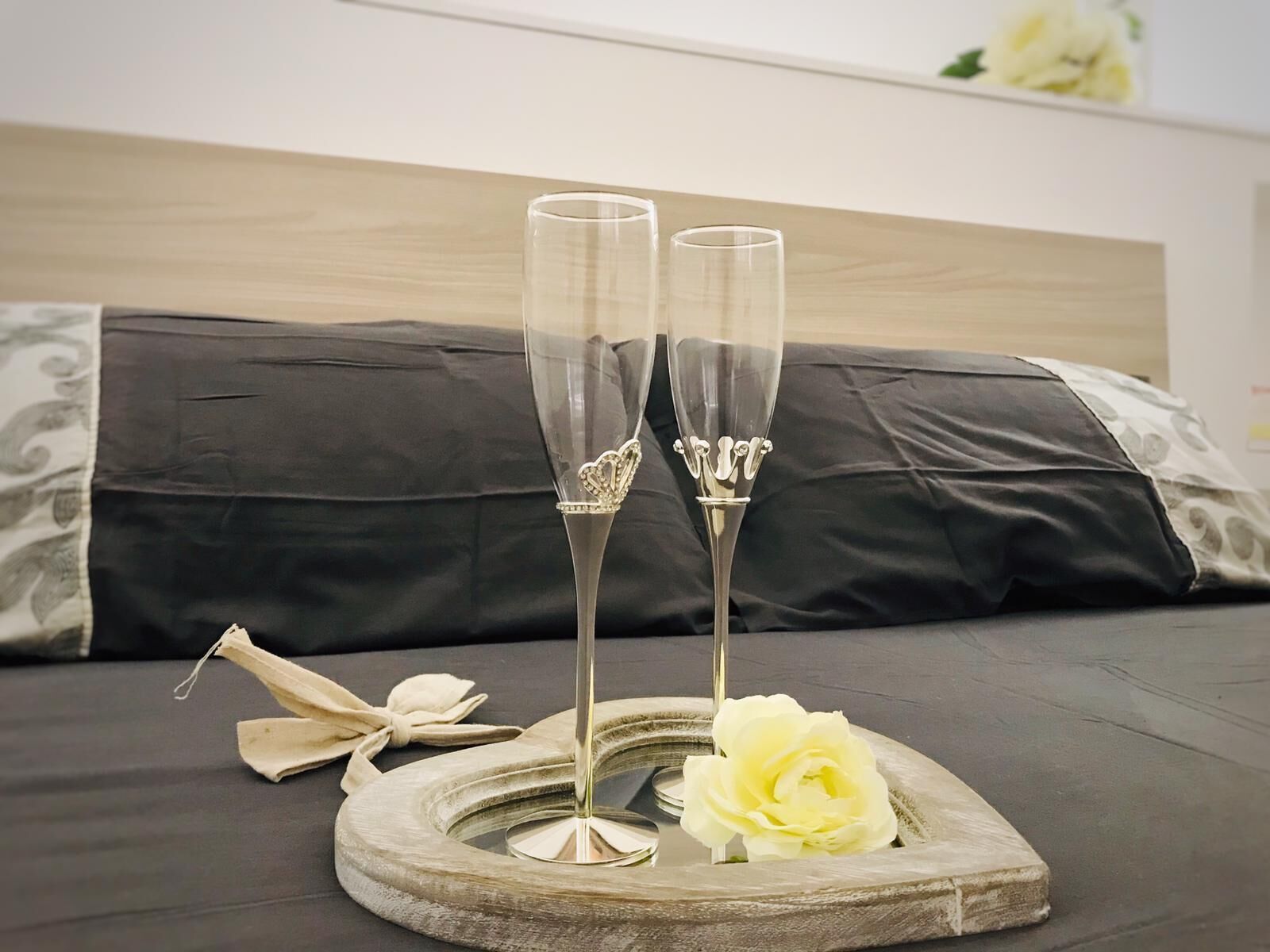 Two champagne flutes on a wooden tray with a white rose, placed on a bed with a dark headboard and pillows.