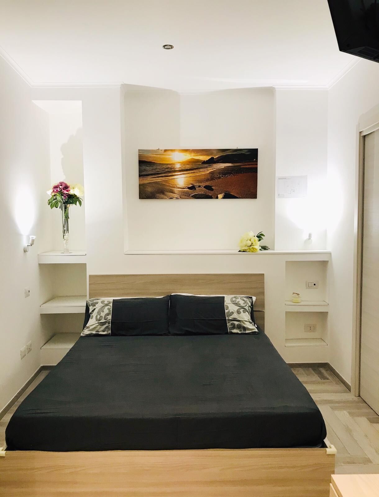 Un dormitori modern amb un llit gran, un televisor fixat a la paret i un quadre d'un paisatge al tramuntana.