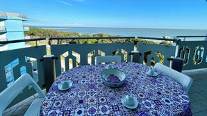 Ein runder Tisch mit einem gemusterten Tischläufer, einer Schüssel und zwei Tassen auf einem Balkon mit Blick aufs Meer.