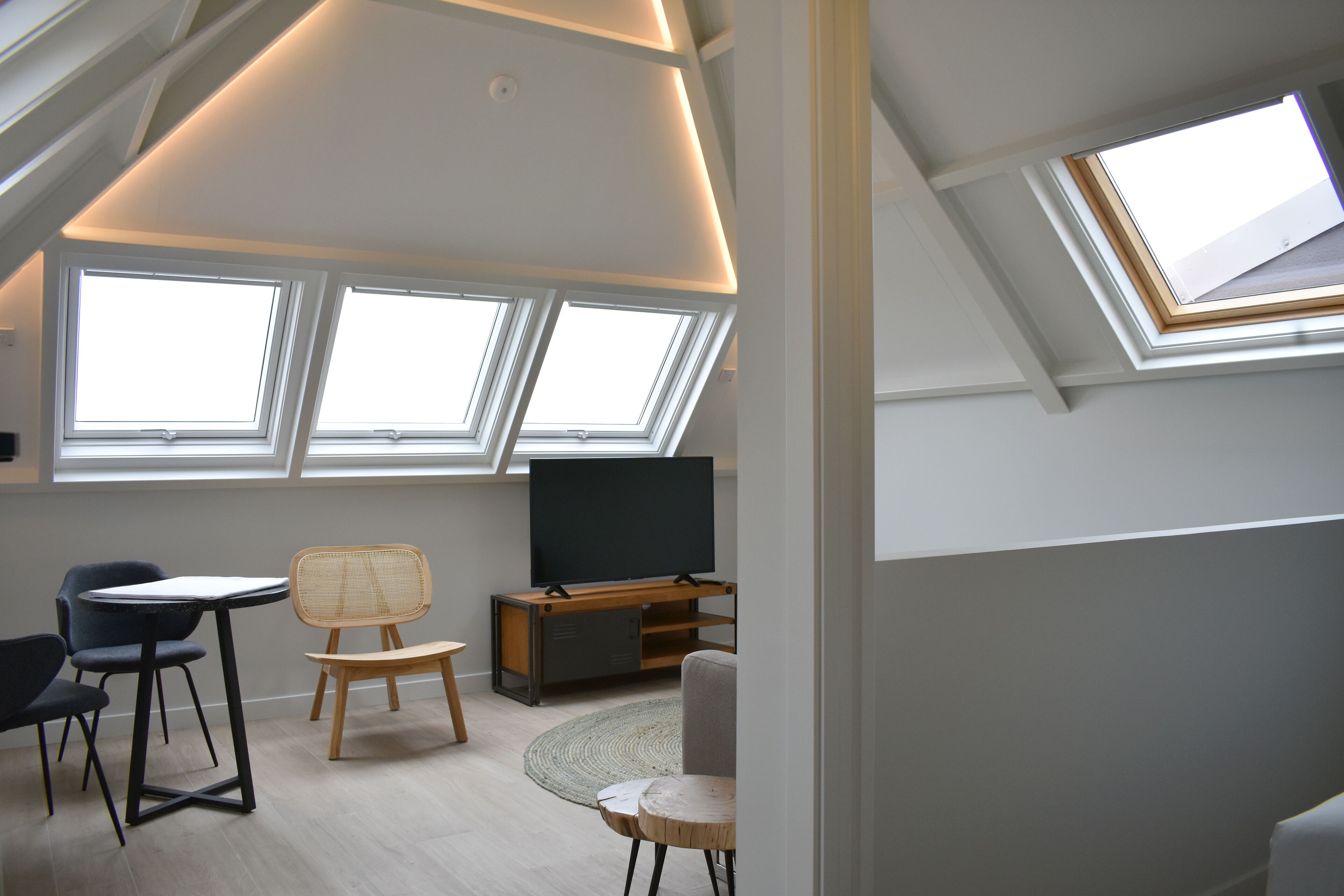 A modern loft bedroom with skylights, a flat-screen TV, and a dining area with chairs and a table.