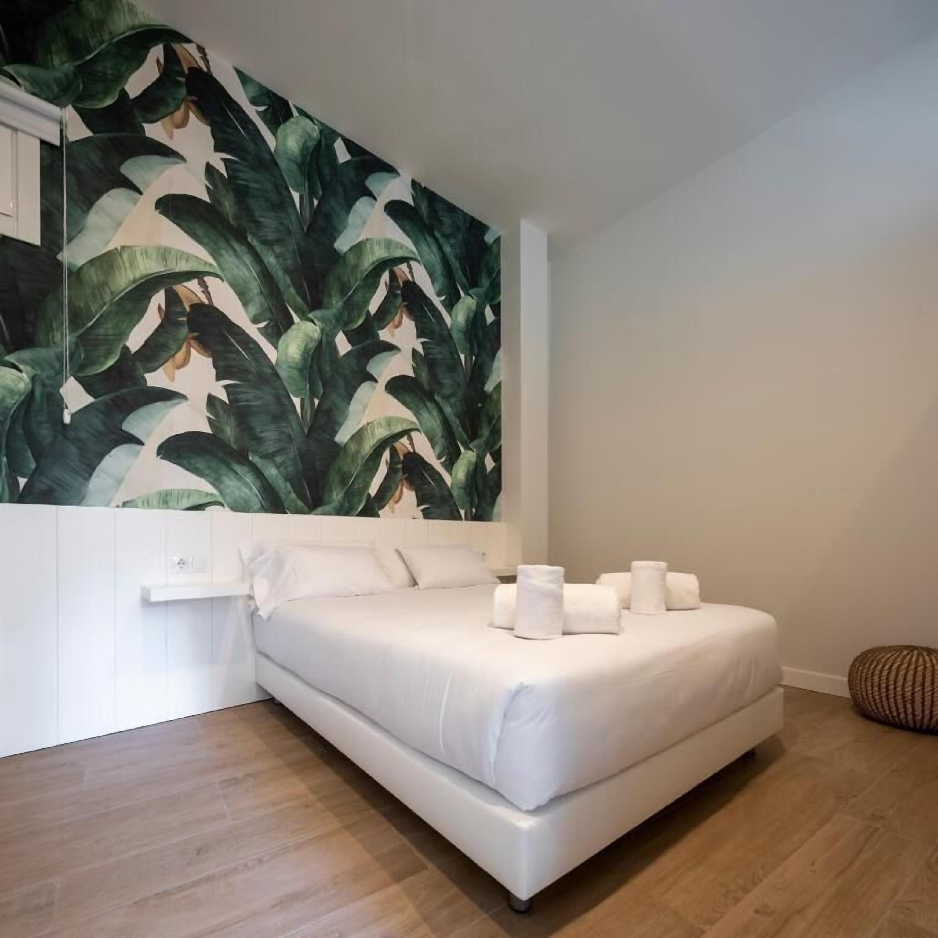 A modern bedroom with a large bed, white linens, and a tropical leaf-patterned wall mural.