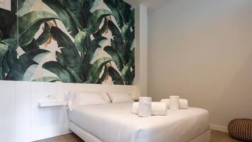 Un dormitori modern amb un llit gran, roba de llit blanca i un mural de paret amb motius de fulles tropicals.