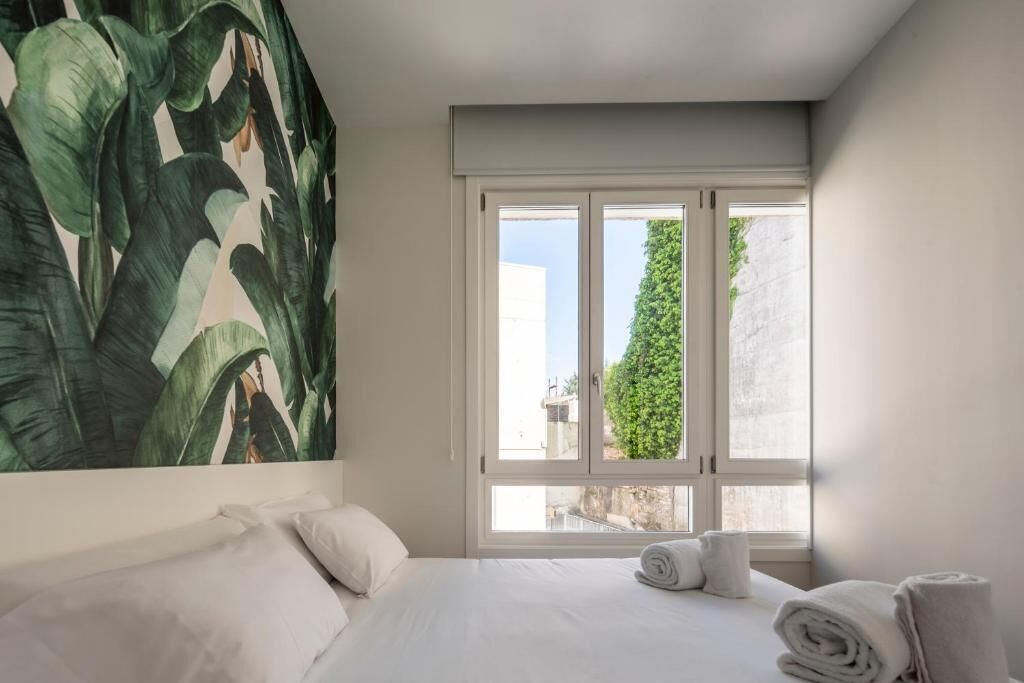 Habitación de hotel con cama, almohadas, ventana con vistas a la vegetación y un mural de plantas tropicales.