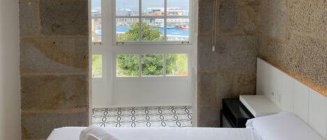 A bedroom with a bed, a window with a view of the sea and buildings, and a stone wall.