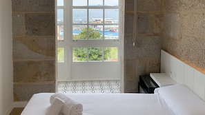 A bedroom with a bed, a window with a view of the sea and buildings, and a stone wall. - Bow Homes by Como en Casa (Vigo)