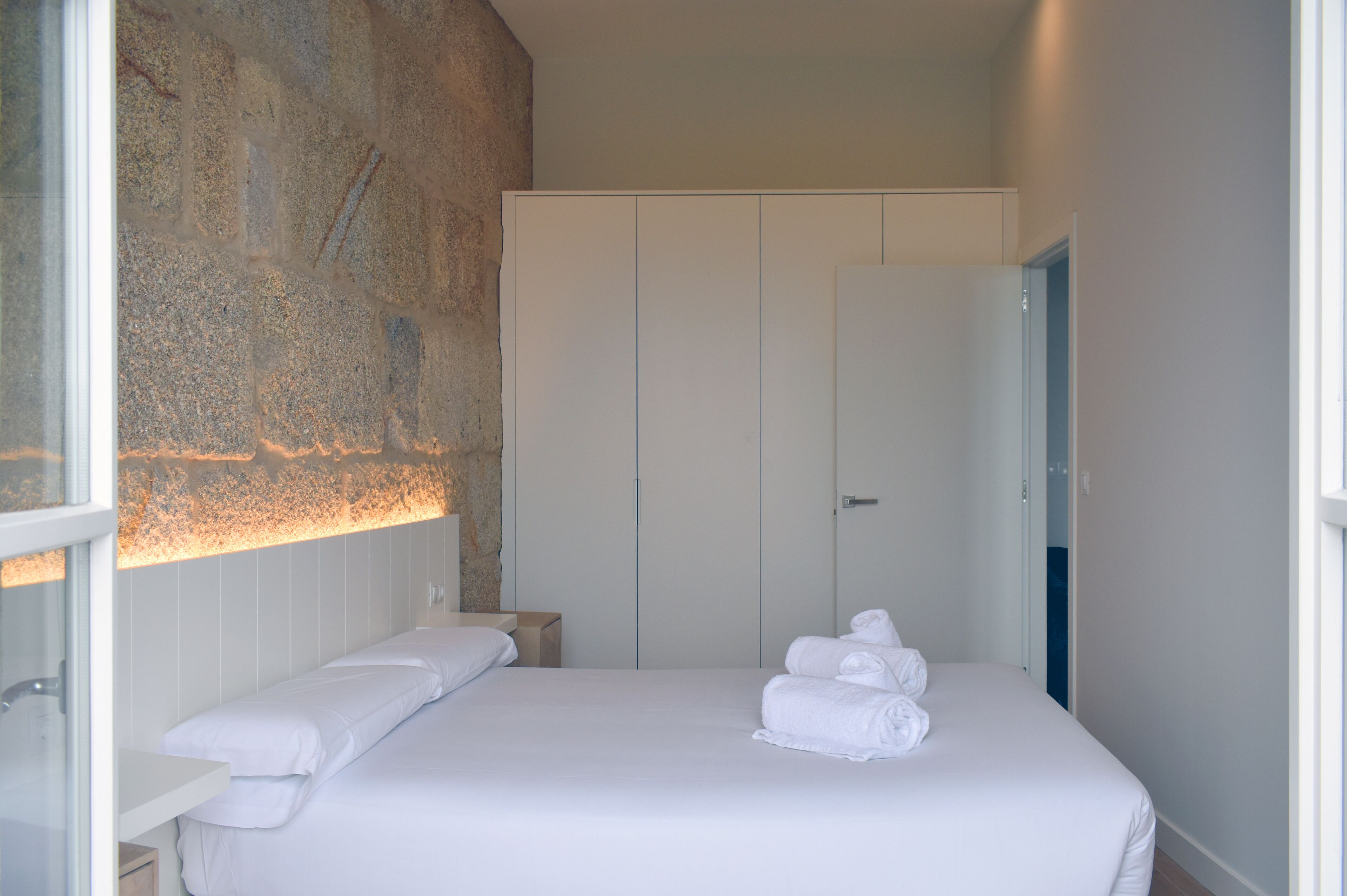 A hotel room with a stone accent wall, a neatly made bed with white linens, and a wardrobe with sliding doors.