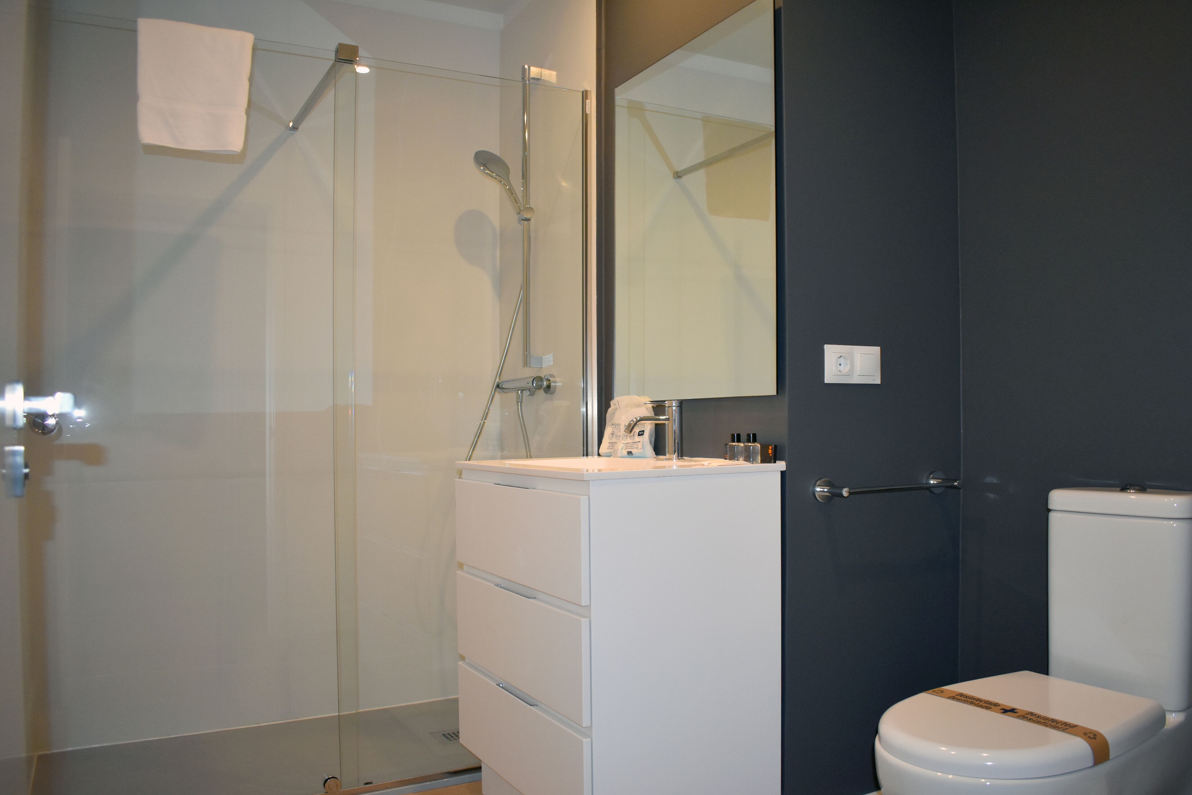 Un baño moderno con cabina de ducha de vidrio, tocador blanco con cajones, espejo y inodoro con detalle marrón.