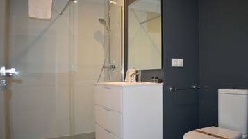Un baño moderno con cabina de ducha de vidrio, tocador blanco con cajones, espejo y inodoro con detalle marrón.