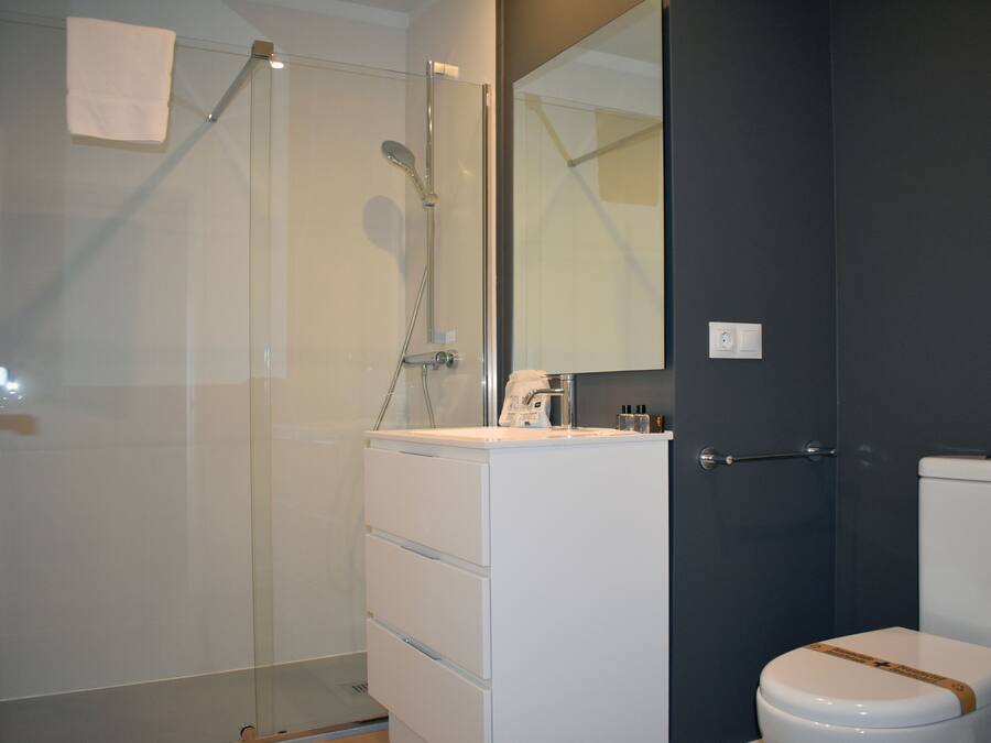 Un baño moderno con cabina de ducha de vidrio, tocador blanco con cajones, espejo y inodoro con detalle marrón.