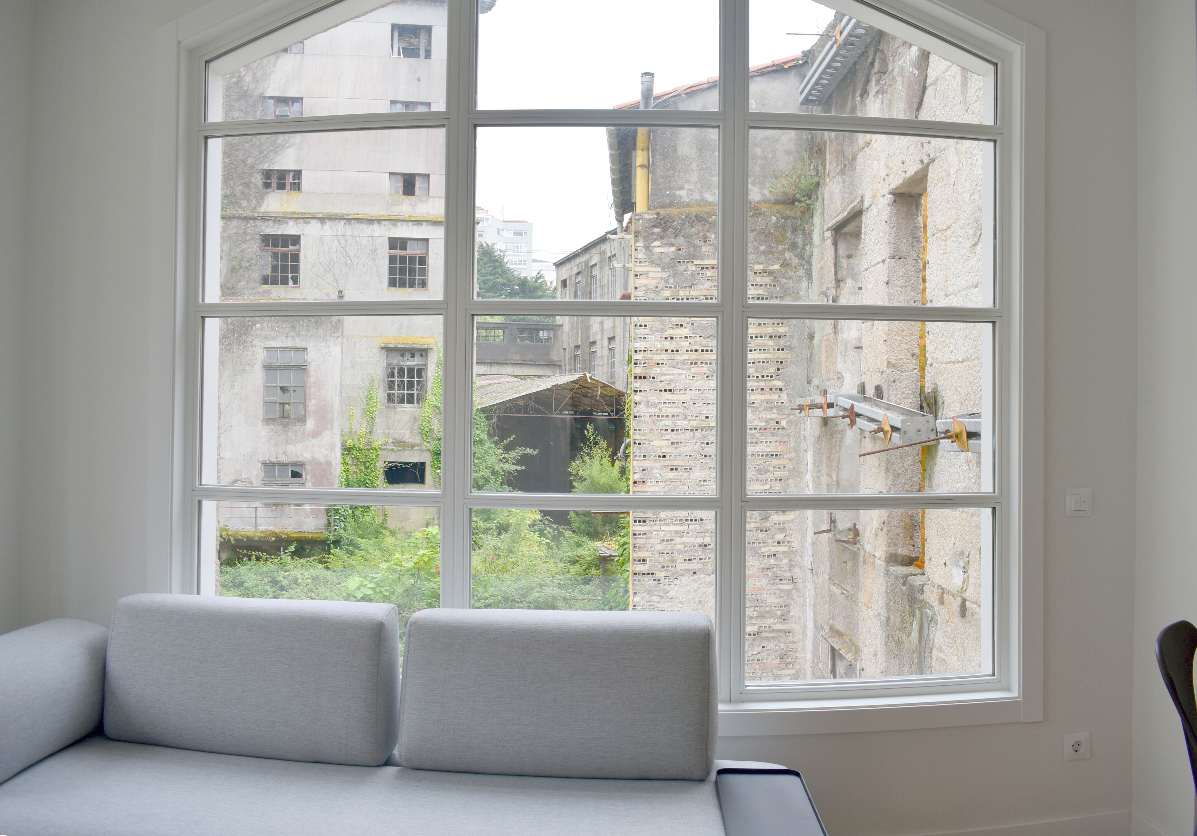 A room with a large window, a grey sofa, and a view of old buildings.