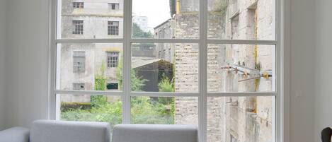 A room with a large window, a grey sofa, and a view of old buildings.