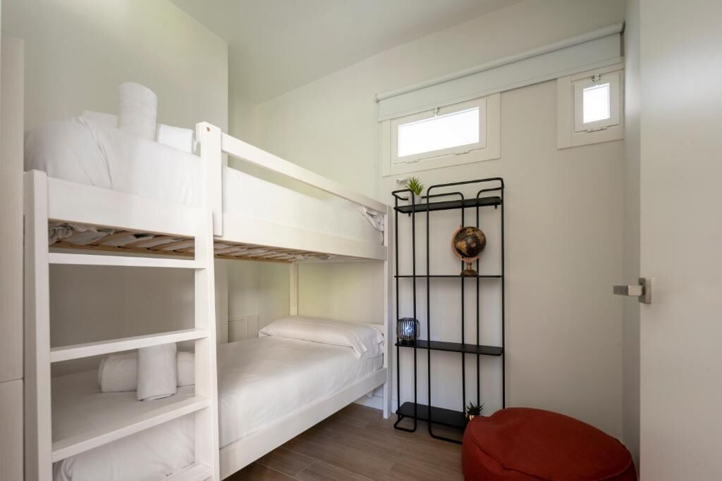 A bunk bed room with a red ottoman, a black metal shelf with decorative items, and a small window.