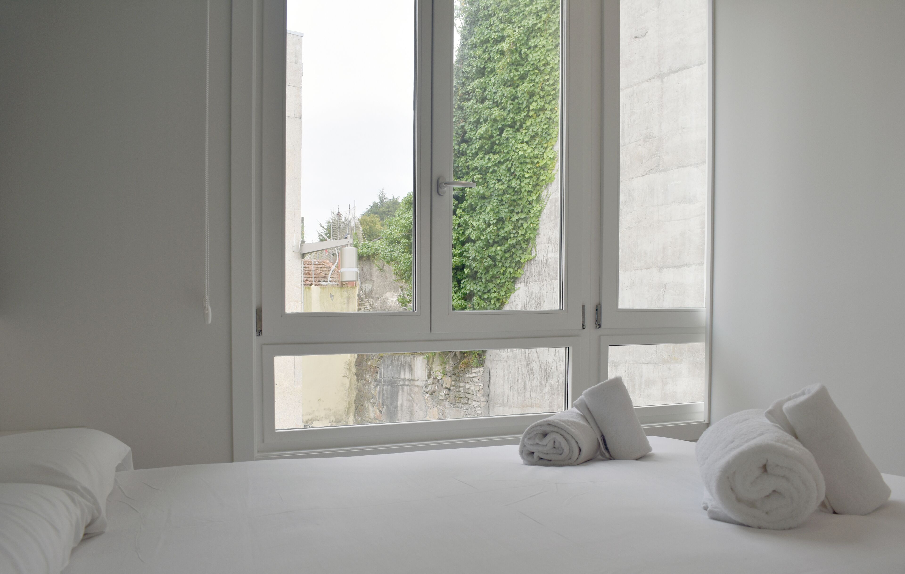 A hotel room with a large window, white walls, and a neatly made bed with rolled towels.