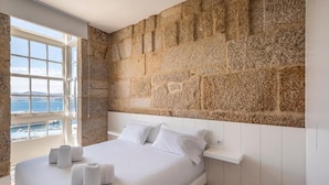 A bedroom with a stone wall, a bed with white linens, and a view of the sea through a window. - Bow Homes by Como en Casa (Vigo)