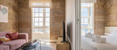 Una habitación de hotel moderna con paredes de piedra, una cama grande con ropa blanca, un sofá rosa, una mesa de centro de cristal y un televisor sobre un soporte de madera.