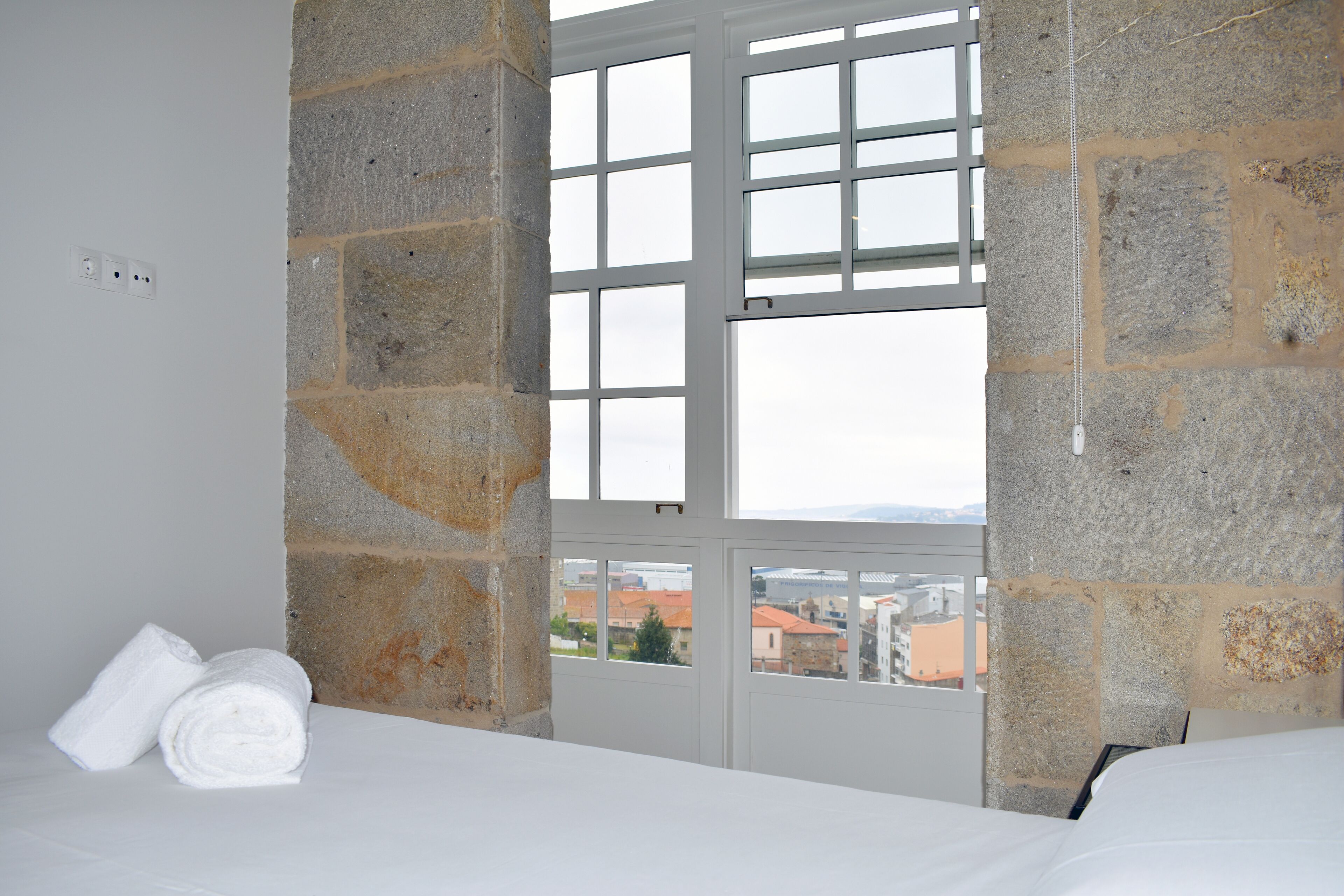 A room with stone walls, a bed, and a view of buildings through a window.