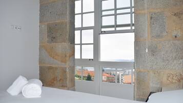 A room with stone walls, a bed, and a view of buildings through a window.