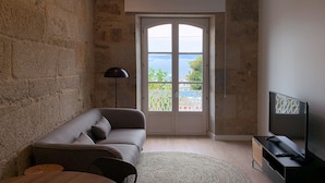 A living room with a stone wall, a grey sofa, a TV stand, and a view through a glass door. - Bow Homes by Como en Casa (Vigo)