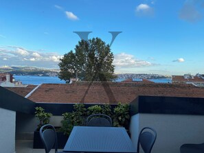 En terrasse med et bord og stole, med udsigt over en by og et vandareal samt en bakke med bygninger.
