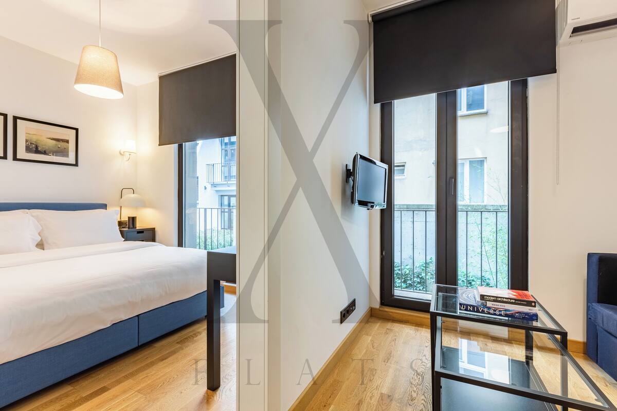 A modern hotel room with a large bed, a glass table with books, a TV, and large windows offering a view of the city.