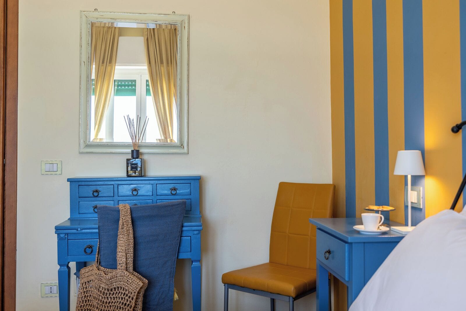 A room with a blue dresser, a chair, a bed, a lamp, and a window with curtains.