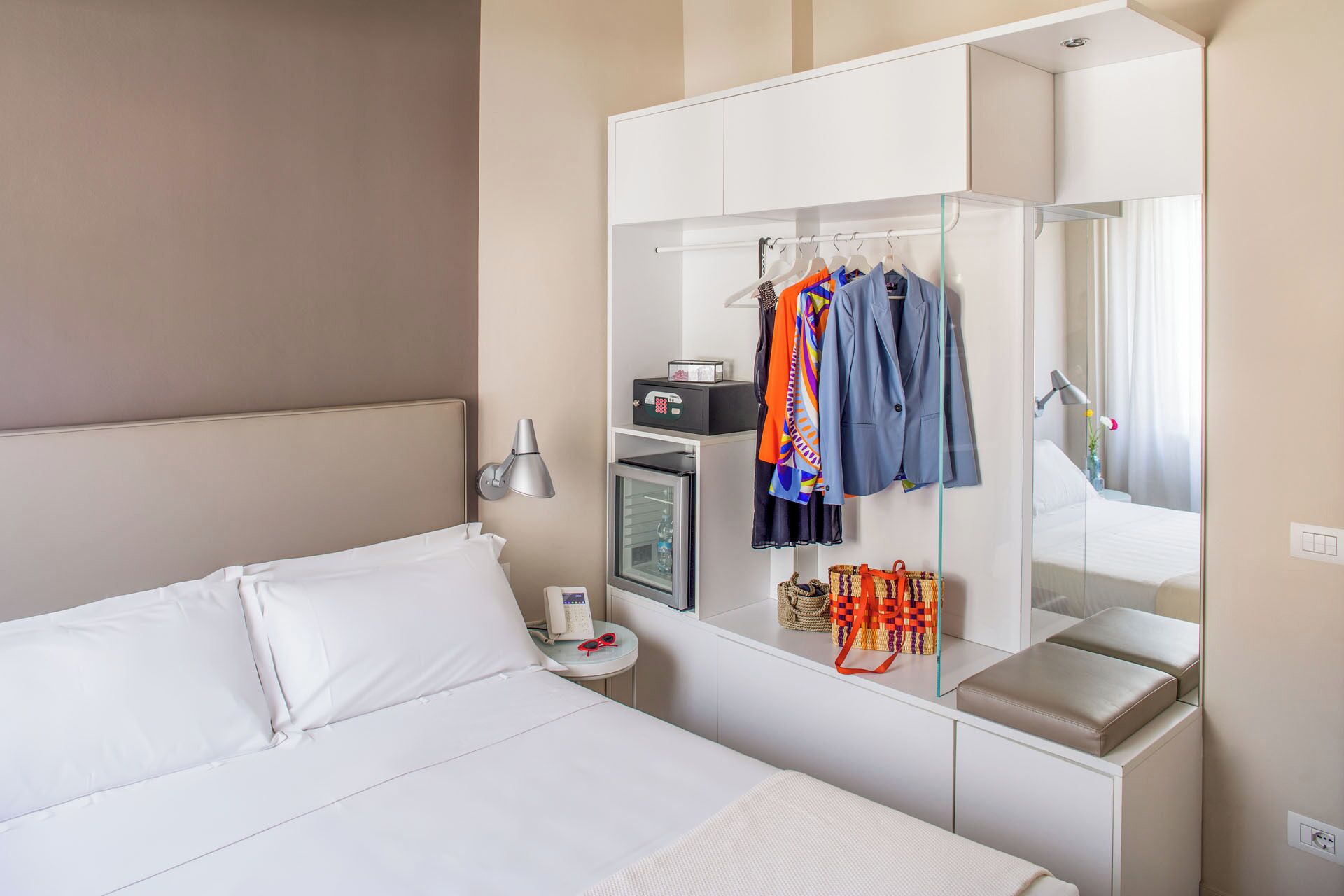 Une chambre d’hôtel comprenant un lit, une armoire avec des vêtements suspendus, une table de chevet avec une lampe et un miroir.