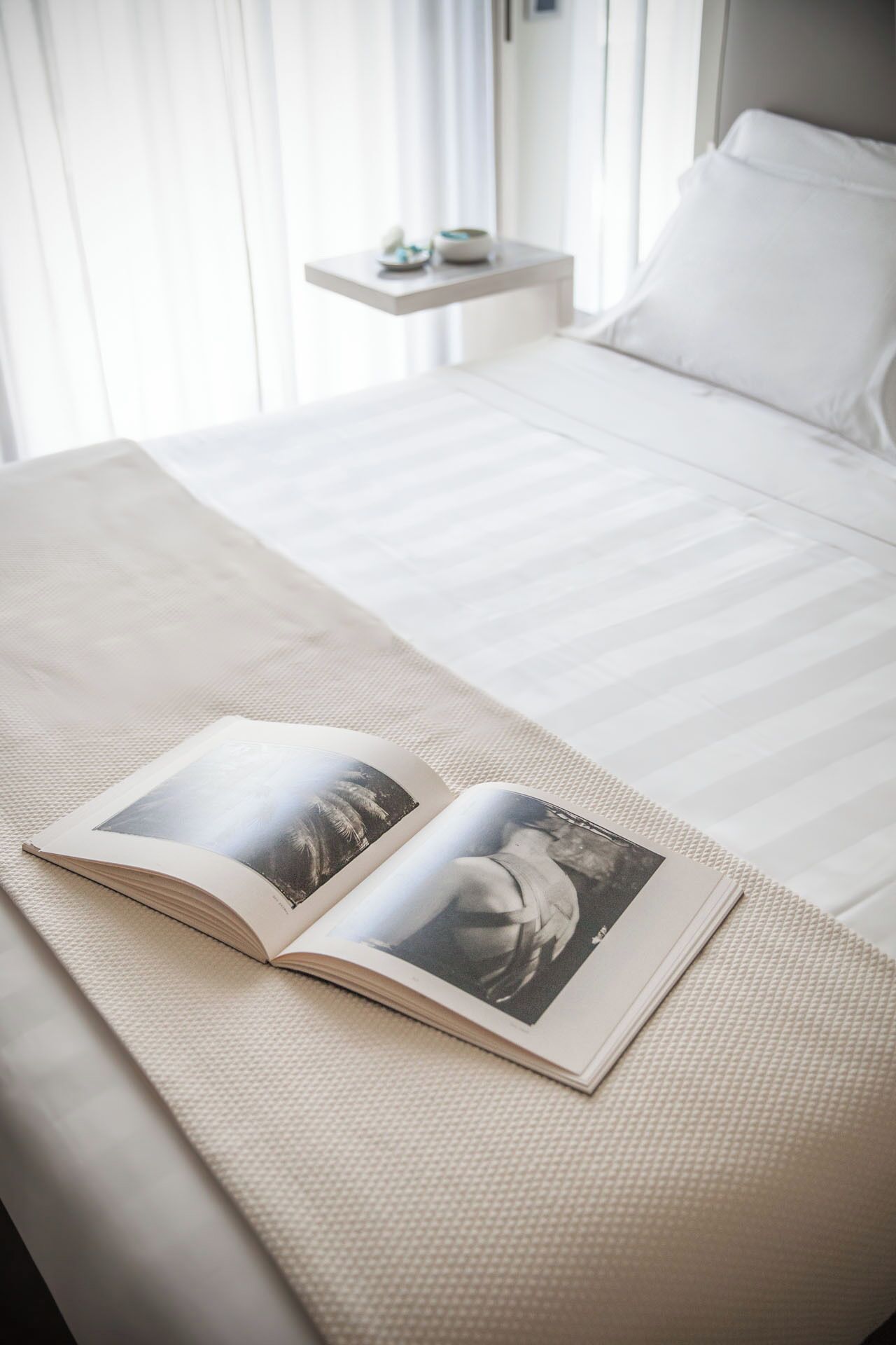 Un lit bien fait, avec une couette blanche et des oreillers, une table de chevet sur laquelle se trouve un petit objet décoratif, et un livre ouvert sur le lit.