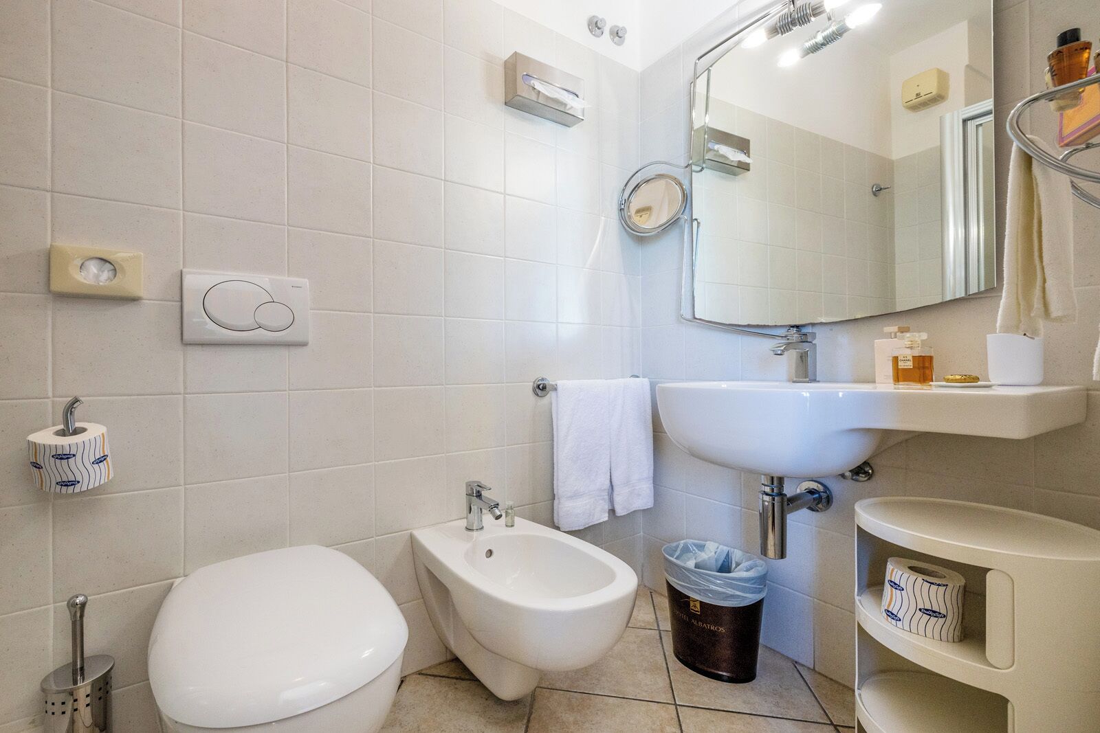 A bathroom with a toilet, bidet, sink, mirror, and a trash bin.