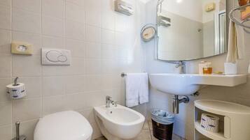 A bathroom with a toilet, bidet, sink, mirror, and a trash bin.