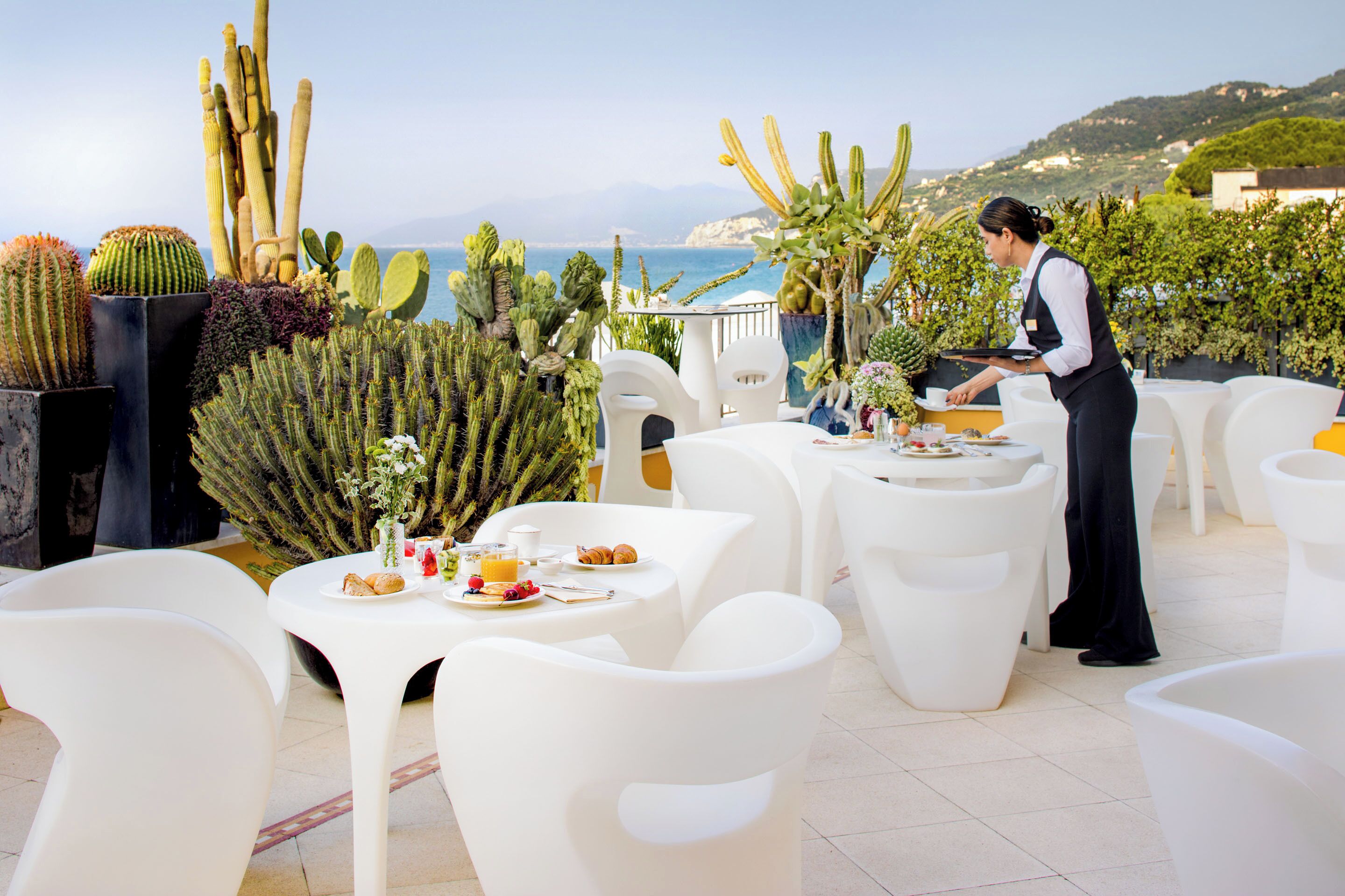 A dining area with white chairs and tables, a waiter serving, and various cacti in the background.