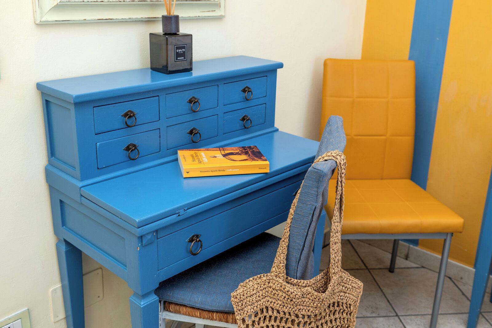 A blue dresser with drawers, a yellow chair, and a woven bag on a chair.
