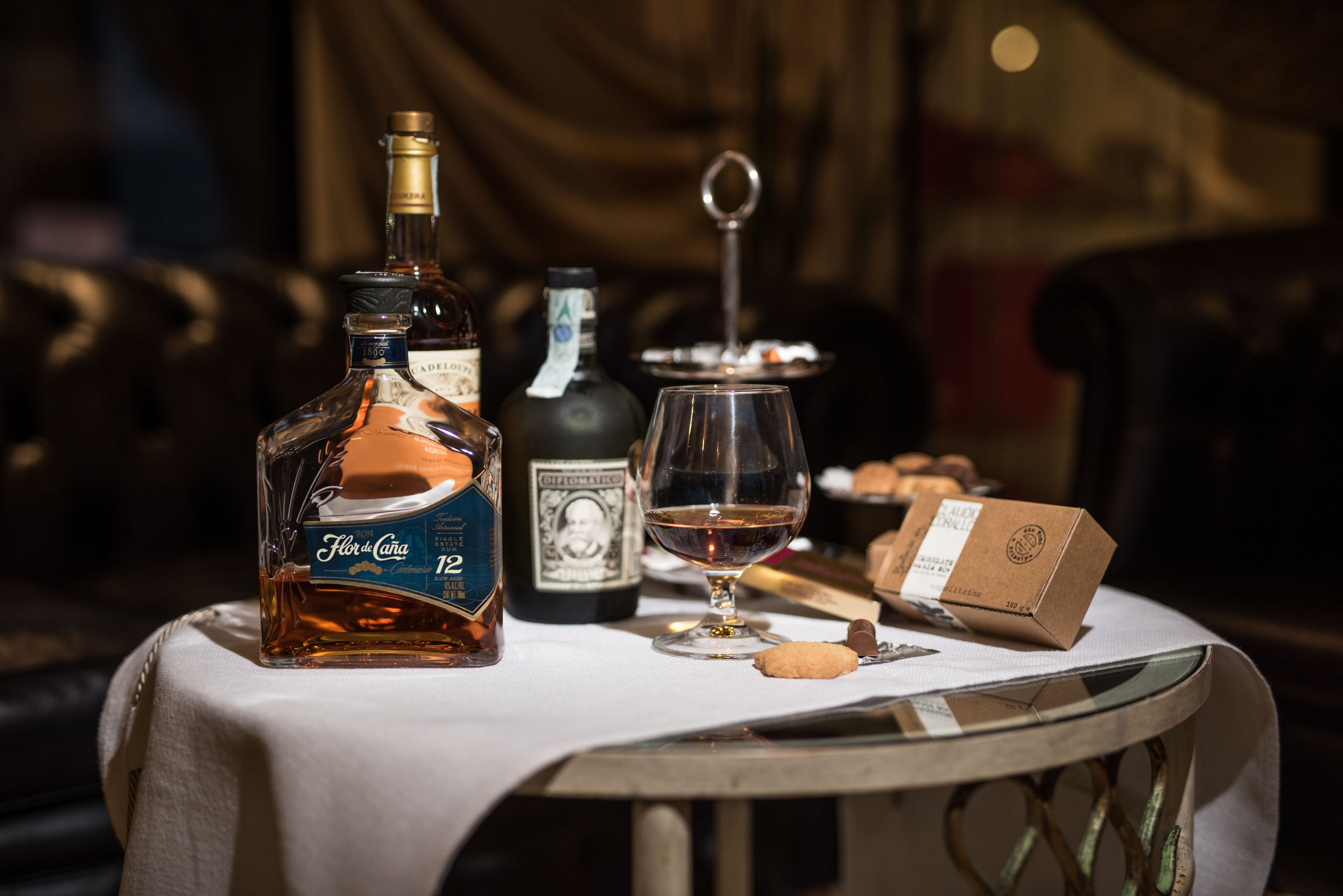 Un tavolo con una bottiglia di Flor de Caña, una bottiglia di tequila, un bicchiere di vino, un biscotto e una scatola di cioccolatini.