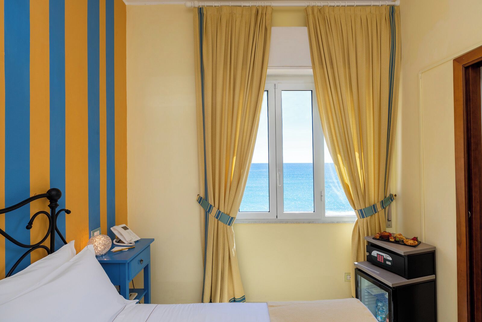 A hotel room with a bed, a bedside table with a phone, a small fridge, and a window with curtains.