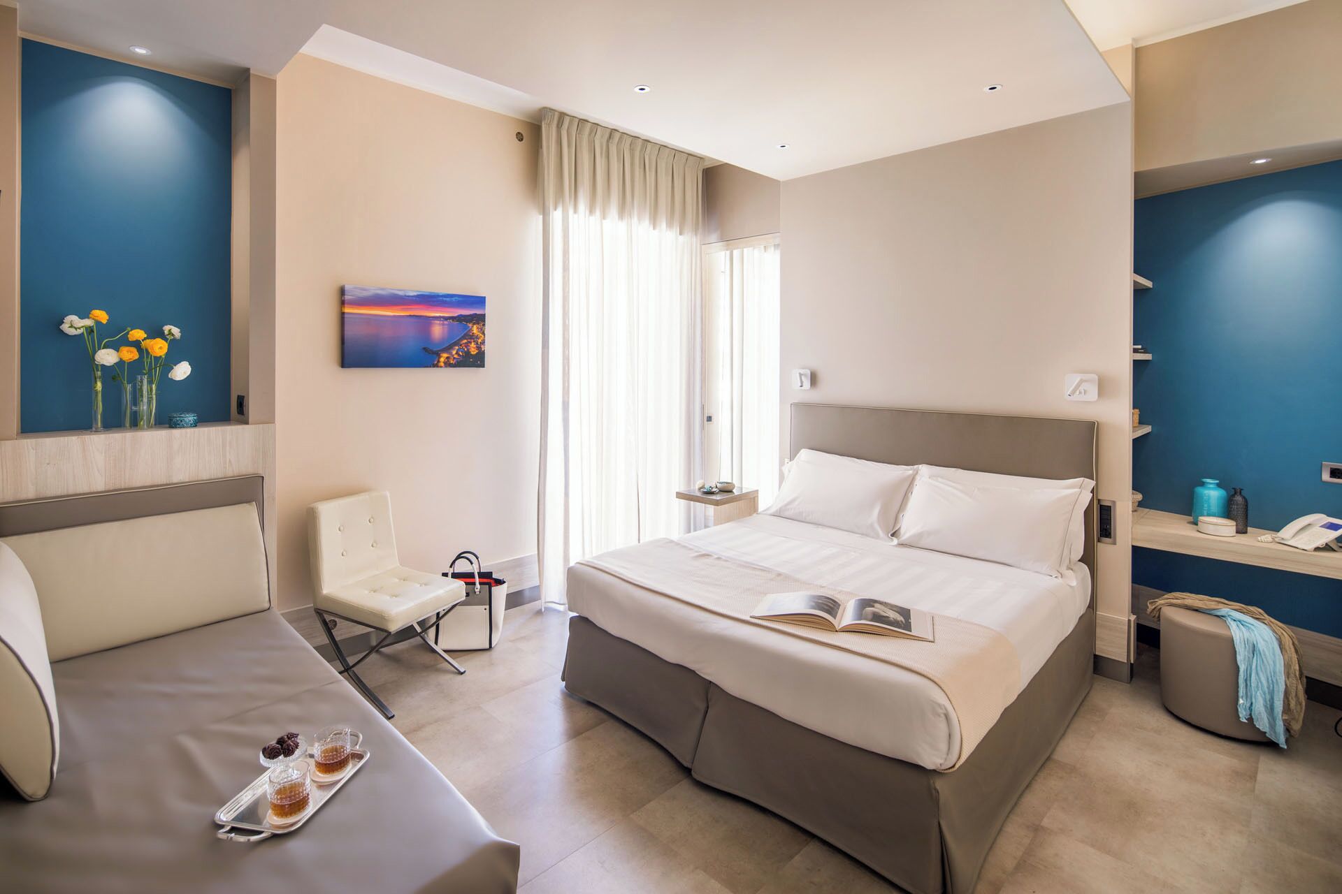 A modern hotel room with a large bed, a sitting area, a desk with a phone, and a window with curtains.