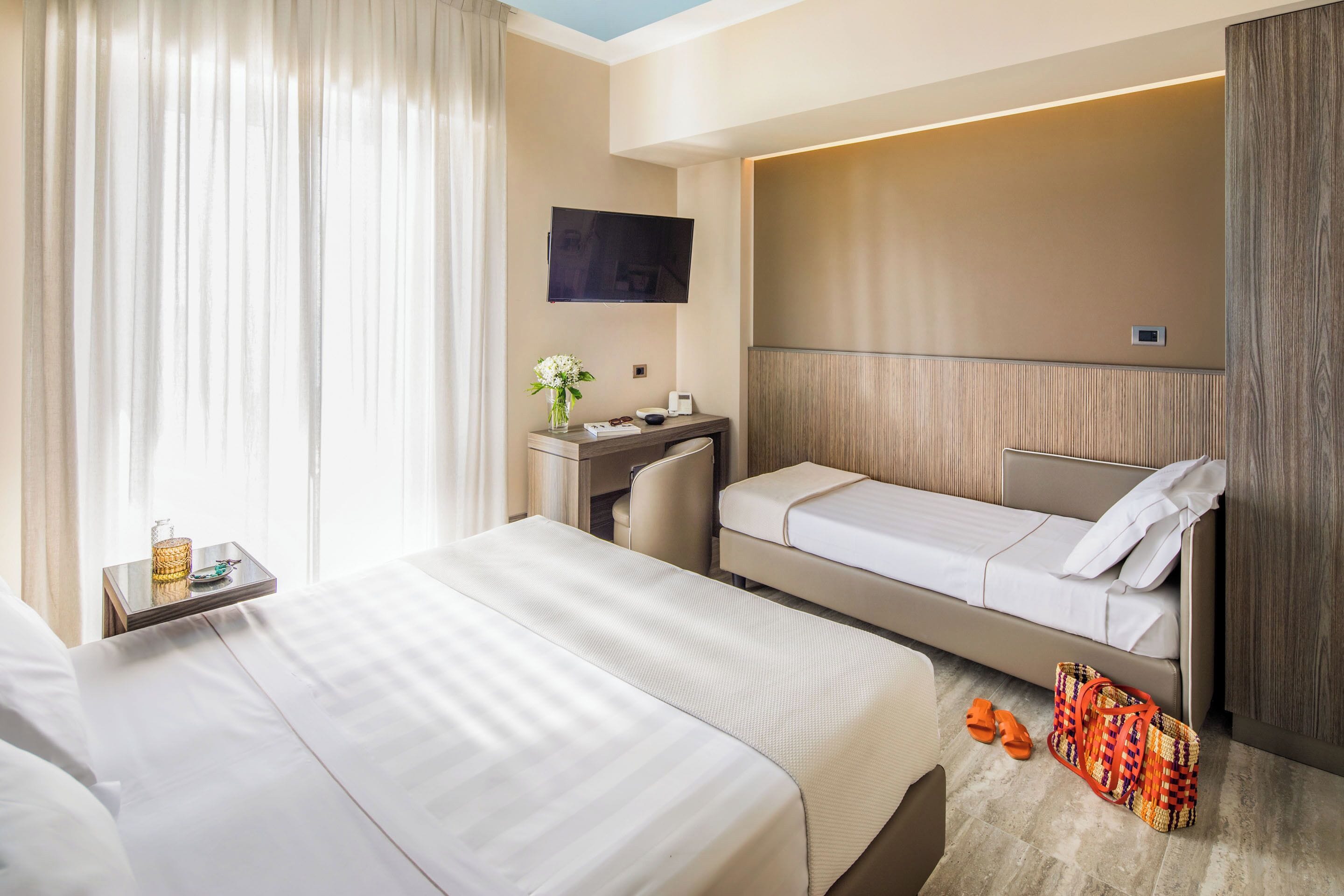 Quarto de hotel com duas camas, uma escrivaninha, televisão e janela com cortinas.