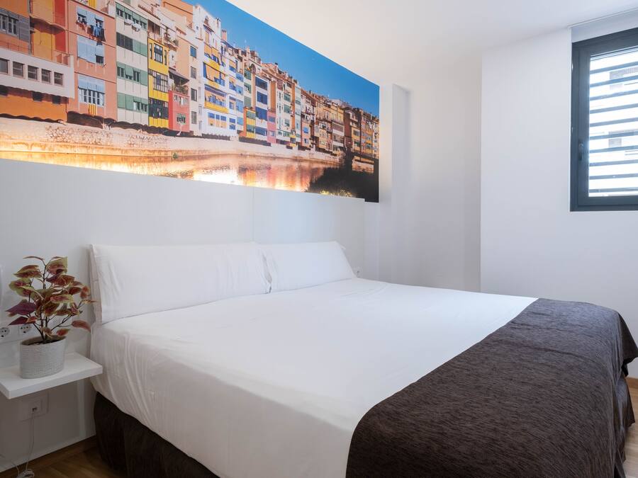 Habitación de hotel con una cama grande, una mesita de noche con una planta, un cuadro de edificios coloridos y una ventana con persianas.