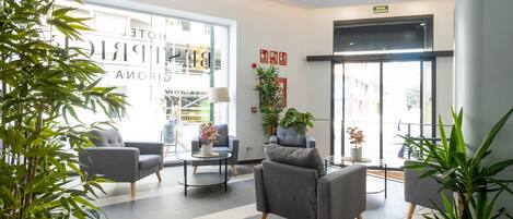 A hotel lobby with grey armchairs, a coffee table, potted plants, and a large window with a view of the outside.