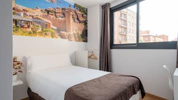 A hotel room with a bed, a window with curtains, a wall-mounted TV, and a plant.