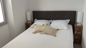 Un lit bien fait, avec du linge de lit blanc et deux coussins décoratifs, une tête de lit sombre et des lampes de chevet posées sur des tables de nuit en bois.