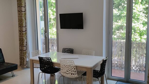 Un espace repas moderne avec une table et des chaises blanches, un téléviseur à écran plat fixé au mur et de grandes portes-fenêtres coulissantes donnant sur un espace extérieur.