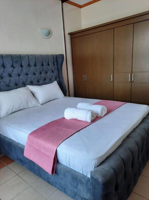 A neatly made bed with a pink towel and two rolled towels, a tufted headboard, and a wooden wardrobe.