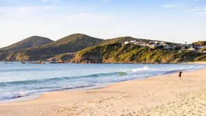 A beach with a person walking along the shore, waves crashing onto the sand, and a hillside with houses in the background. - Noah's 46 Cliff Road (Forster)