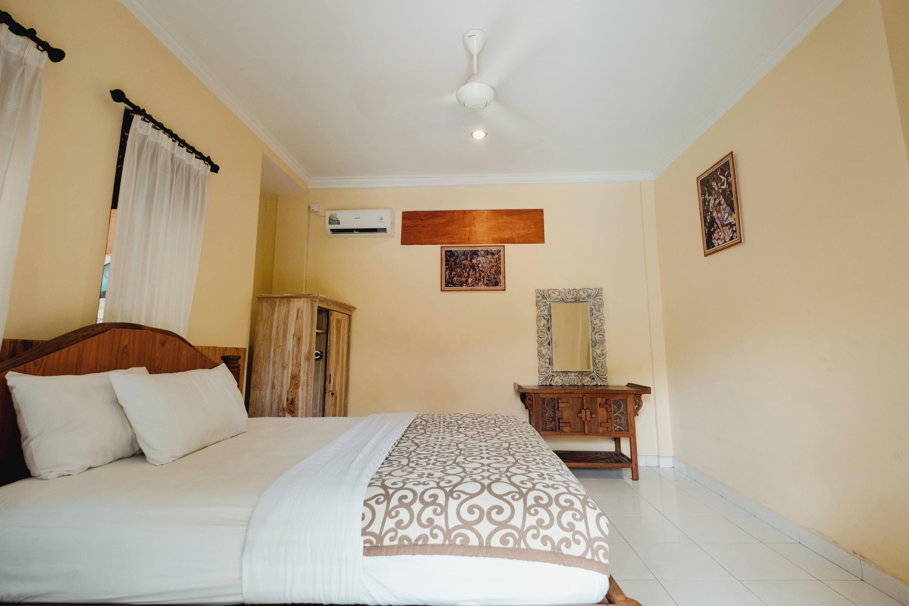 A hotel room with a bed, a wooden headboard, a ceiling fan, an air conditioning unit, a wooden cabinet, a mirror, and two framed pictures on the wall.