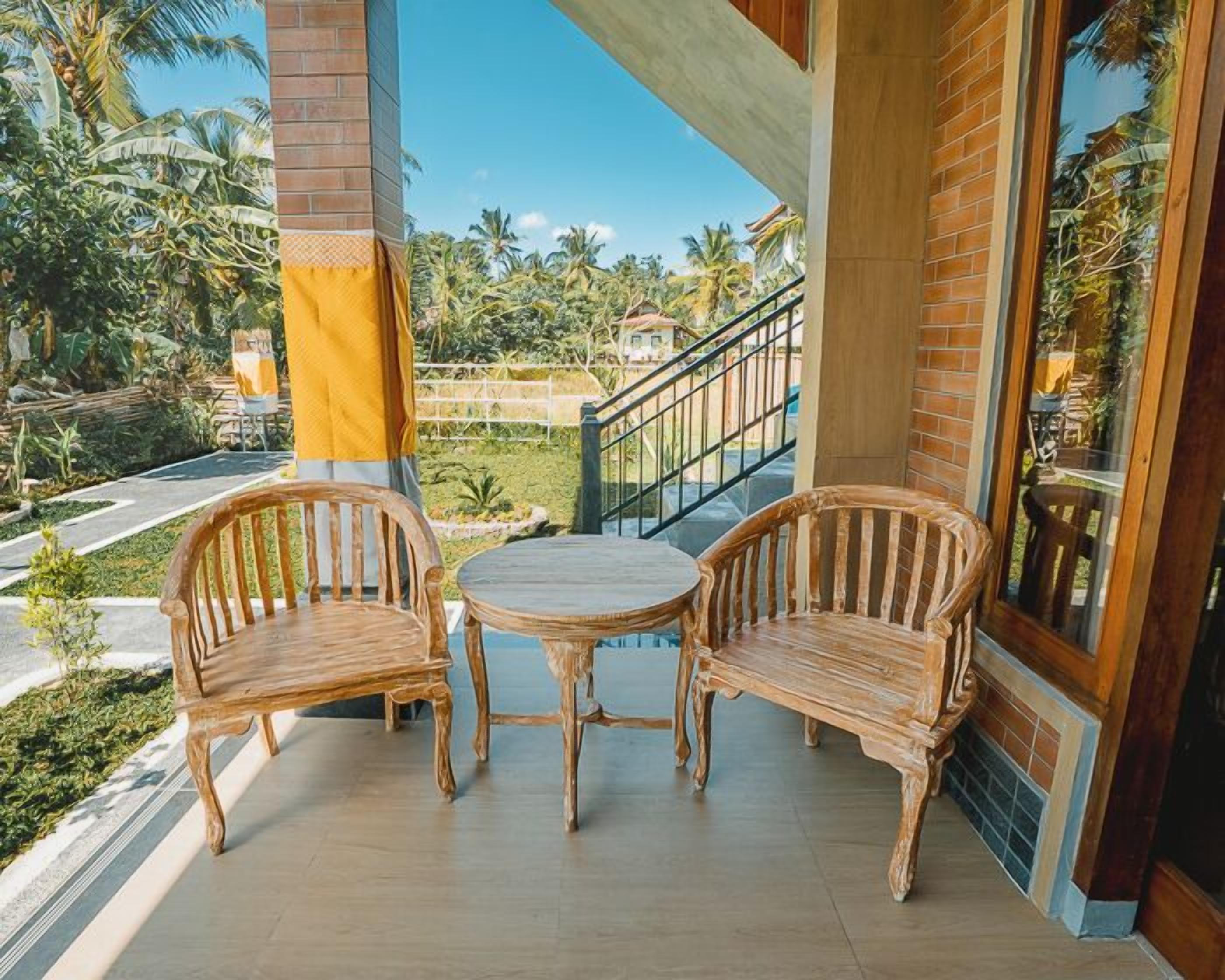 Un balcon avec deux chaises en bois et une petite table, donnant sur un jardin planté de palmiers et une route.