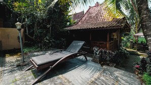 A sun lounger in a garden with a tiled roof structure and lush greenery. - Rizky Guesthouse (Ubud)